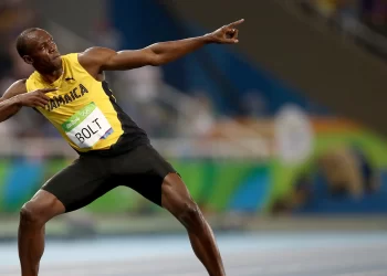 A sprinter in a yellow and black uniform with "Jamaica" and "BOLT" visible on his chest, Usain Bolt strikes a celebratory pose, pointing upward with one arm while the other is bent across his body. The background shows a blurred track and audience. | FintechZoom