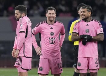 Three soccer players wearing pink jerseys, standing on a soccer field. The middle player is smiling while the other two have neutral expressions. A referee, dressed in yellow, is seen in the background. The jerseys feature black accents and a team emblem. | FintechZoom