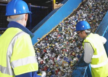 Two workers wearing blue helmets and reflective vests are observing and sorting aluminum cans on a conveyor belt in a recycling facility. The conveyor belt, part of an advanced waste management system, is filled with numerous crushed cans moving upwards for processing. | FintechZoom