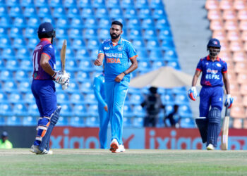 A cricket player in a blue India uniform is seen celebrating on the pitch. Two batsmen in blue and red uniforms, playing for the Nepal National Cricket Team, are standing at either end of the pitch. The stadium's stands in the background feature empty blue and orange seats, capturing a moment in the ongoing Nepal National Cricket Team vs India National Cricket Team timeline. | FintechZoom