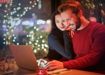 A couple sits at a cozy, dimly lit cafe table, smiling and looking at a laptop. The man types on the keyboard while the woman rests her head on his shoulder. Warm, twinkling lights and festive decorations are visible in the background as they explore mortgage online options together. | FintechZoom