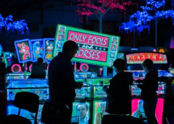 People in front of illuminated arcade game machines with colorful signs in a dimly lit room. A prominent sign reads "ONLY FOOLS and HORSES." Neon lights in blue and pink hang from the ceiling, casting a vibrant glow on the scene, as someone eagerly approaches the Jackpot Slots. | FintechZoom