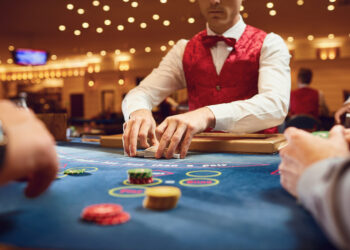 A casino dealer in a red vest and bow tie efficiently shuffles cards at a poker table adorned with colorful chips. The warmly lit casino setting has other players partially visible, all focused on the dealer's productive actions. | FintechZoom