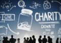 Silhouettes of people standing and chatting in front of a blue background filled with various hand-drawn philanthropy-themed illustrations, including a jar with coins, the words "GIVE" and "CHARITY," and icons like a piggy bank, heart, puzzle piece, and medical symbol. | FintechZoom