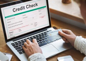 A person is holding a credit card and using a laptop on a wooden table. The laptop screen displays a credit check form with various fields including personal details, status, and income, integral for effective Credit Monitoring. The person appears to be filling out the form. | FintechZoom
