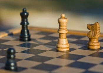 A close-up of a chessboard features three prominent chess pieces: a black king on the edge, a wooden king in the center, and a wooden knight nearby. Another black pawn is slightly visible in the lower corner. The chessboard, illuminated by natural light outdoors, plays out like strategic moves managing credit risks. | FintechZoom