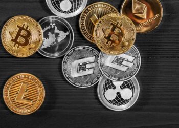 A collection of cryptocurrency coins, including Bitcoin, Ethereum, Litecoin, NEO, and Ripple, scattered on a dark wooden surface. The cryptocurrency coins are in various colors, predominantly gold and silver, with distinct symbols and designs representing different digital currencies. | FintechZoom