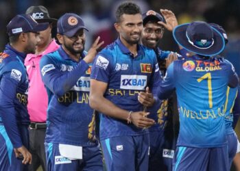 A group of Sri Lankan cricket players, dressed in blue and yellow uniforms, is gathered on the field celebrating a moment during an India vs Sri Lanka match. One player is central, being congratulated by teammates who are smiling and clapping. An umpire stands in the background. | FintechZoom