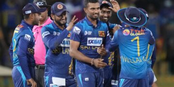 A group of Sri Lankan cricket players, dressed in blue and yellow uniforms, is gathered on the field celebrating a moment during an India vs Sri Lanka match. One player is central, being congratulated by teammates who are smiling and clapping. An umpire stands in the background. | FintechZoom