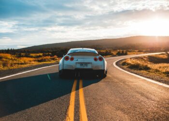 A white sports car with red tail lights glides down a winding, empty road during sunset. The surrounding landscape consists of grassy fields and distant hills under a partly cloudy sky. The sunlight casts a warm glow over the scene, reminding drivers to consider car insurance for peace of mind. | FintechZoom