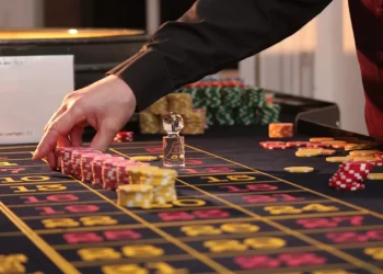A person's hand is arranging stacks of poker chips on a roulette table, indicating a casino setting. The roulette wheel is partially visible on the left side of the image, with various colored chips and betting grid numbers on the table, evoking the thrill found in online gambling experiences. | FintechZoom