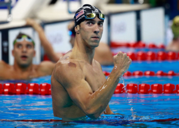 A male swimmer wearing a swim cap and goggles stands in the pool, raising his fist in a celebratory gesture. He appears victorious. In the background, another swimmer looks on. Lane dividers are visible in the water, evoking an Olympic atmosphere. | FintechZoom