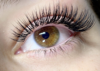 A close-up of an eye with a light brown iris showcasing long, curled, and voluminous eyelashes enhanced by lash extensions. The surrounding skin is smooth, and the lighting highlights the well-defined lashes. | FintechZoom