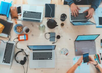 A top-down view of a cluttered table with five laptops, each being used by different people. The table also features notebooks, headphones, a teapot, a cereal bowl, a phone, water bottles, and various other items. This scene suggests an IT staffing collaborative work environment. | FintechZoom