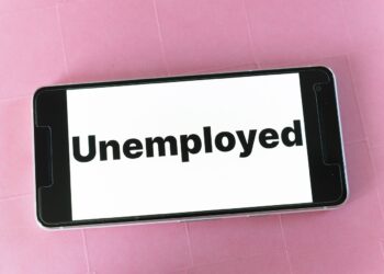A smartphone lies on a pink surface with the words "Unemployment Protection" displayed in bold black text on its screen. | FintechZoom