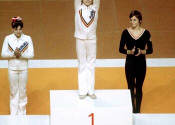 Three gymnasts stand on a podium. The gymnast in the center is on the highest step, raising her arms like Nadia Comăneci. The gymnast on the left is in second place, clapping, while the gymnast on the right, in fourth place, is applauding. The podium is labeled 2, 1, and 3. | FintechZoom