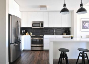Modern kitchen with white cabinets, stainless steel appliances including a microwave, and a dark backsplash. Two black pendant lights hang over a white island with two black stools. The floor is wood. A framed picture hangs on the right wall. | FintechZoom