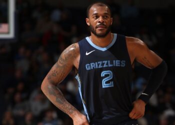 A basketball player, Xavier Tillman, is wearing a dark blue "Grizzlies" jersey with the number 2 printed on it. He has tattoos on his arms and is standing with his hands on his hips on a basketball court, with a blurred crowd in the background. | FintechZoom