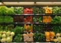 A supermarket grocery produce section displaying an array of fresh vegetables: leafy greens in the top left, red and yellow bell peppers, zucchinis, yellow squash, cucumbers, eggplants, cauliflowers, and pumpkins neatly arranged on multiple shelves. | FintechZoom