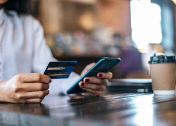 A person holding a credit card in their left hand and a smartphone in their right, likely interacting with a payment processor. They are seated at a wooden table with a takeaway coffee cup nearby. The background is blurred, indicating a coffee shop or similar setting. | FintechZoom