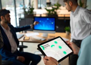 Three people are in a modern office. One person in a blue suit points at a computer screen displaying an analytical dashboard graph. Another person in white stands nearby. A third person holds a tablet showing a "financial planning" dashboard with charts and graphs. | FintechZoom