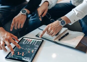 Two people, wearing smart casual attire and watches, are sitting at a table discussing data on a tablet displaying stock market graphs. Open notebooks and a pen indicate a collaborative financial analysis session focused on the best crypto trading bots. | FintechZoom