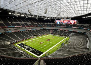 An empty, well-lit football stadium with a translucent arched roof. The green field features yard lines and end zones with the name “Allegiant” on one side. Large video screens at either end display team logos and scores. Rows of empty seats surround the field, awaiting the many ways to watch Super Bowl 2024. | FintechZoom