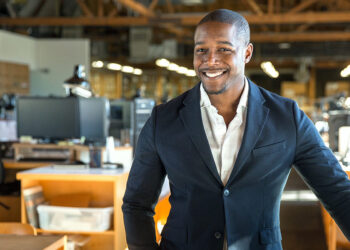 A man wearing a dark blazer and white shirt is smiling and standing in an office environment with multiple desks, computer monitors, and lights hanging from the ceiling. The background features large windows and an exposed wooden beam ceiling, resembling a modern daycare business headquarters. | FintechZoom