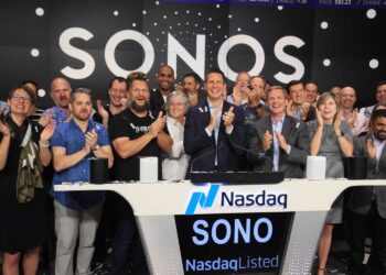 A large group of people stand on a stage under a sign that reads "SONOS", clapping and smiling. At the center, a person stands at a podium with the Nasdaq logo, celebrating an event related to Sonos stock. Electronic devices are also visible on the podium. | FintechZoom