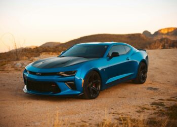 A blue sports car is parked on a dirt road in a desert landscape during sunset. The car features a sleek design with black accents and is positioned at an angle, showcasing its front and side profile. Rocky hills and a clear sky are visible in the background—perfect for those searching to find the best car insurance. | FintechZoom