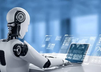 A robot with a sleek white and metallic design is seated at a desk, typing on a futuristic transparent tablet. The background is a blurred, blue-toned room filled with abstract digital interfaces, suggesting a high-tech environment buzzing with venture capital innovation. | FintechZoom