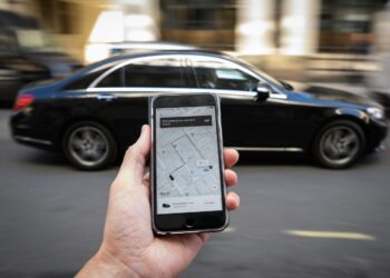 A person holding a smartphone with a ride-hailing app open, showing a map with a car icon. In the background, a black car is in motion on a city street, creating a blurred effect due to the movement. The scene suggests organizing an Uber ride as news of Uber stock surges. | FintechZoom
