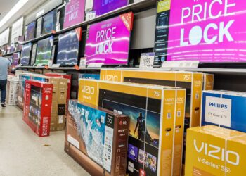 A store aisle filled with large screen TVs in boxes, including Vizio, LG, and Philips. Overhead displays announce "PRICE LOCK" promotions. Two people are seen from the back walking down the aisle, exploring the extensive Vizio stock available. | FintechZoom