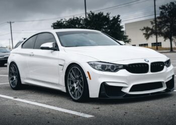 A sleek white BMW car, potentially attractive to car insurance companies, is parked in a nearly empty parking lot on a cloudy day. The car has a sporty design with large alloy wheels and dark-tinted windows. Trees and buildings are visible in the background. | FintechZoom
