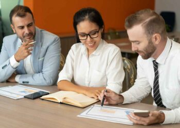 Three people are at a meeting in a conference room. The woman in the middle, an insurance broker, is smiling and looking at a notebook, while the man on the right is pointing at a document and talking. The man on the left is thoughtfully observing the discussion. | FintechZoom
