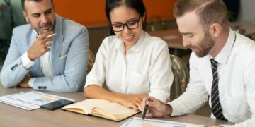 Three people are at a meeting in a conference room. The woman in the middle, an insurance broker, is smiling and looking at a notebook, while the man on the right is pointing at a document and talking. The man on the left is thoughtfully observing the discussion. | FintechZoom