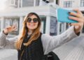 A person wearing sunglasses, a knit hat, and a gray sweater takes a selfie with a blue smartphone. They are smiling and making a peace sign with their left hand. The background shows a city street with buildings and sunlight filtering through, perhaps perfect for those looking to buy Instagram likes. | FintechZoom