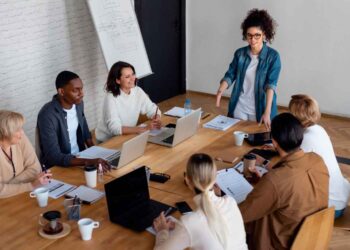 A diverse group of people are gathered around a wooden table for a meeting. Some have laptops open, while others have notebooks to jot down key points. A person standing and gesturing appears to be leading the discussion on various benefits. There's a whiteboard in the background with diagrams. | FintechZoom