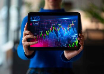 A person in a blue shirt holds a tablet displaying a colorful stock market graph with various line charts, numbers, and grid lines, indicative of fluctuating stock prices. The image subtly highlights the sophisticated interface typical of fintech apps. The background is blurred and indistinct. | FintechZoom