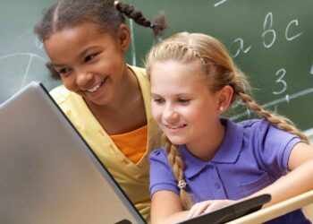 Two young girls are sitting at a desk in a classroom, smiling and looking at a laptop together, possibly checking their Blooket login. The girl on the left has braided hair and is wearing a yellow shirt, while the girl on the right has blonde hair in a braid and is wearing a purple shirt. A chalkboard is in the background. | FintechZoom