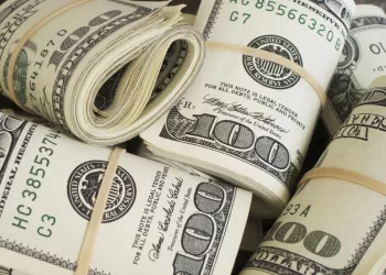 Bundles of U.S. hundred-dollar bills are secured with rubber bands, exemplifying meticulous bankroll management. The stacks are rolled and folded, displaying the intricate details of the currency, including the portrait of Benjamin Franklin and various serial numbers. | FintechZoom