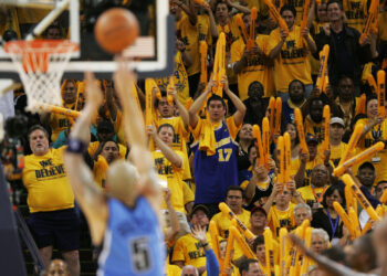 A basketball player wearing a blue away uniform shoots a free throw as fans in yellow "We Believe" shirts cheer and wave orange thundersticks in the stands, creating an atmosphere of Home Court Advantage. The crowd is enthusiastic and focused on the game action. | FintechZoom