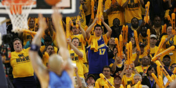 A basketball player wearing a blue away uniform shoots a free throw as fans in yellow "We Believe" shirts cheer and wave orange thundersticks in the stands, creating an atmosphere of Home Court Advantage. The crowd is enthusiastic and focused on the game action. | FintechZoom