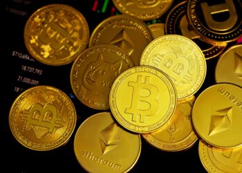 A pile of various gold-colored cryptocurrency coins, including Bitcoin, Ethereum, and Dogecoin, is spread out over a background of a digital screen displaying financial data and charts. The coins feature iconic symbols representing each cryptocurrency token. | FintechZoom