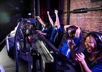 A team of four people in matching blue jerseys sits at a row of computers in an esports gaming setup. They are wearing headsets, and three of them are cheering energetically, while one has their hands raised in celebration. They seem to have won a game. | FintechZoom
