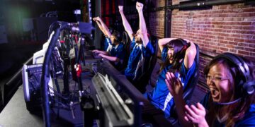 A team of four people in matching blue jerseys sits at a row of computers in an esports gaming setup. They are wearing headsets, and three of them are cheering energetically, while one has their hands raised in celebration. They seem to have won a game. | FintechZoom