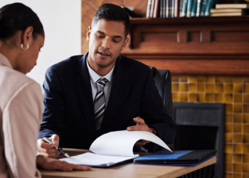 A man in a suit is seated at a table, holding a document and engaging in a discussion with a woman who is also seated and writing. They appear to be in a professional setting, likely discussing legal matters, with books and a fireplace in the background. | FintechZoom