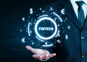 A person in a suit extends their hand, projecting a holographic image featuring the word "FINTECH MARKETING" surrounded by various currency symbols ($, €, ¥, £) on a dark background. The image signifies advancements and innovation in financial technology. | FintechZoom