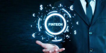 A person in a suit extends their hand, projecting a holographic image featuring the word "FINTECH MARKETING" surrounded by various currency symbols ($, €, ¥, £) on a dark background. The image signifies advancements and innovation in financial technology. | FintechZoom