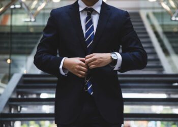 A person dressed in a dark business suit with a blue and white striped tie, possibly the General Counsel, is adjusting their jacket. They are standing in front of a modern stairway in a spacious atrium or lobby, with glass railings and a sleek design. Their face is not visible in the image. | FintechZoom